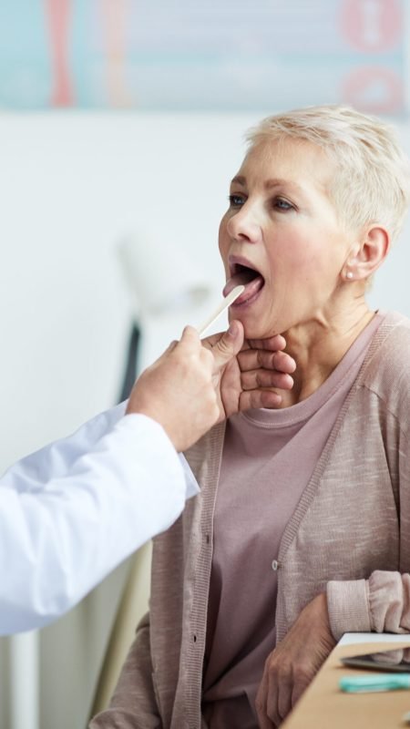 Serious aged male doctor in lab coat using tongue depressor while checking throat of mature lady during medical exam